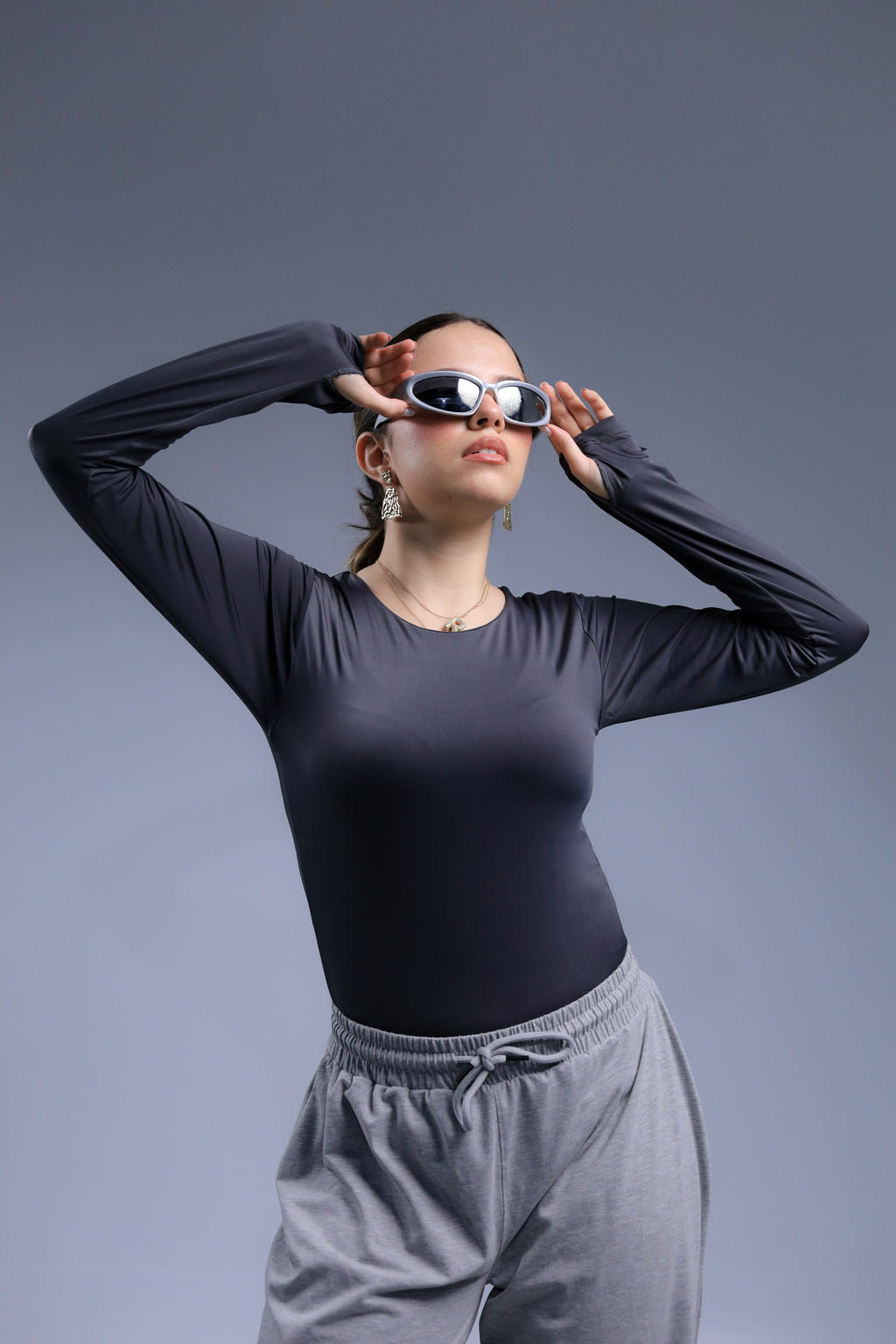 Person wearing a black long-sleeve top and gray pants, adjusting sunglasses against a gray background