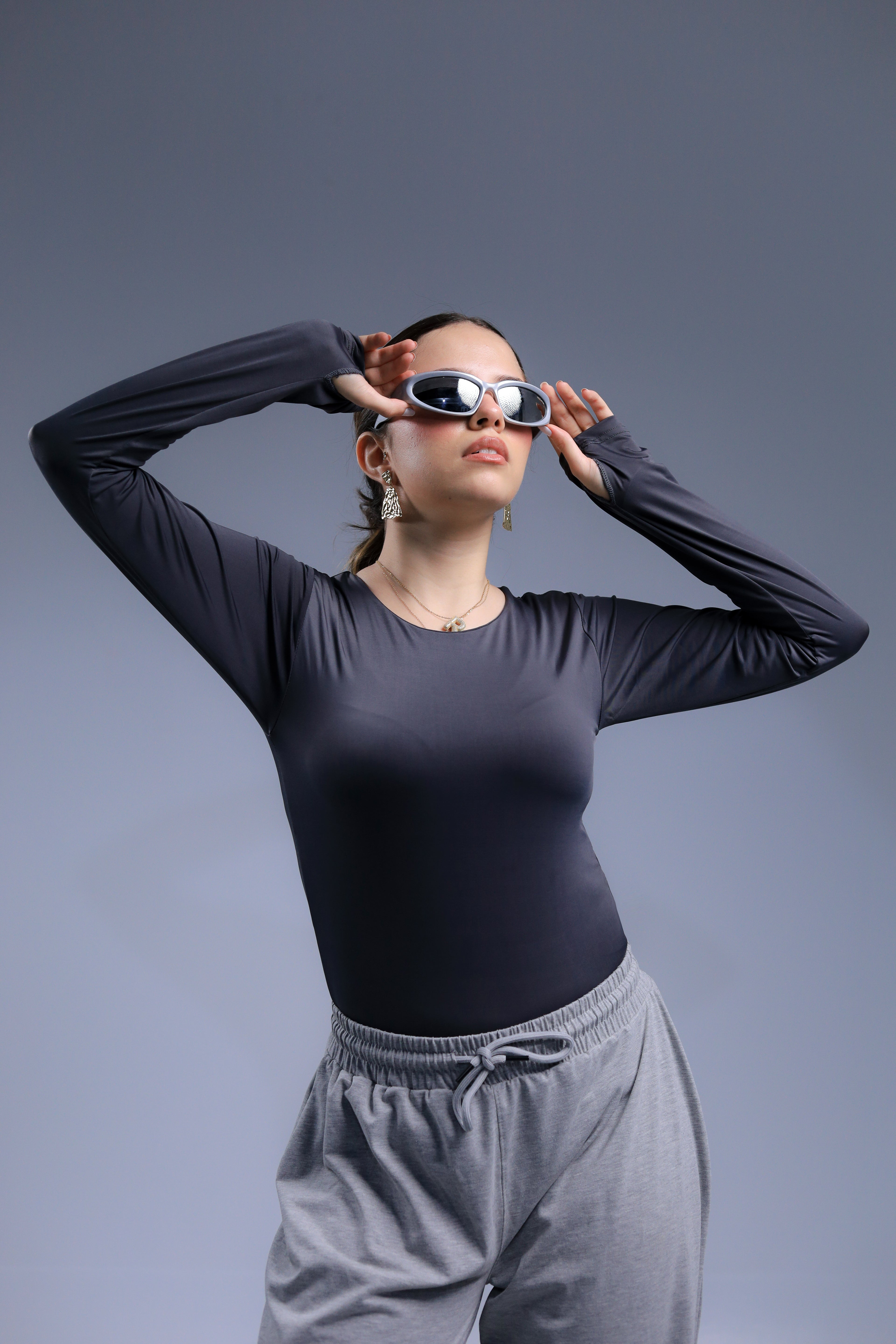 Person wearing a black long-sleeve top and gray pants, adjusting sunglasses against a gray background